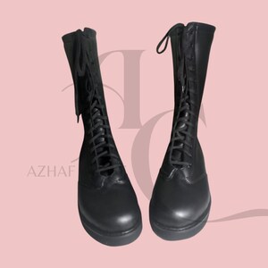 K&ouml;nnte beinhalten: Ein Paar schwarze, kniehohe Stiefel mit Schn&uuml;rsenkeln und Rei&szlig;verschl&uuml;ssen. Die Stiefel sind aus glattem Material gefertigt und haben eine abgerundete Spitze. Der Hintergrund ist einfarbig hellrosa. Das Wort "AZHAF" ist unten links sichtbar.