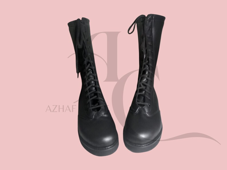 K&ouml;nnte beinhalten: Ein Paar schwarze, kniehohe Stiefel mit Schn&uuml;rsenkeln und Rei&szlig;verschl&uuml;ssen. Die Stiefel sind aus glattem Material gefertigt und haben eine abgerundete Spitze. Der Hintergrund ist einfarbig hellrosa. Das Wort "AZHAF" ist unten links sichtbar.