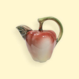 May include: A ceramic jug in the shape of a red apple, featuring a green handle and stem. The jug has a glossy finish and a leaf detail near the spout. This apple-shaped jug is a decorative kitchen item.