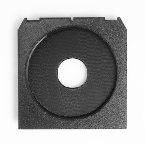 May include: A black, square-shaped object with a circular indentation in the center. The object has a textured surface and a smaller, circular hole in the middle. The design includes two small rectangular cutouts on opposite sides.