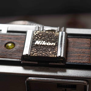 May include: Close-up of a vintage Nikon camera. The camera features a textured, dark brown and gold metal plate with the Nikon logo. The camera body is a mix of dark brown wood and silver metal, with a small yellow circle on the side.