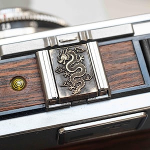 May include: Close-up of a camera with a decorative metal plate featuring a detailed dragon design. The plate is set against a backdrop of dark wood and silver metal components. The camera's lens and other parts are visible.
