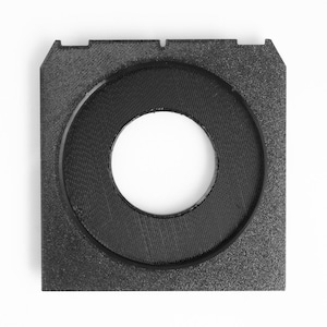 May include: A black, square-shaped 3D-printed object with a circular opening in the center. The object has a textured surface and a raised circular rim around the opening. The corners of the square have small cutouts.