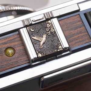 Puede incluir: Primer plano de un detalle de cámara de metal y madera. El punto focal es una placa rectangular con un diseño de colibrí, una luna creciente y estrellas. La placa está enmarcada en plata y sobre un fondo oscuro y texturizado con detalles florales.