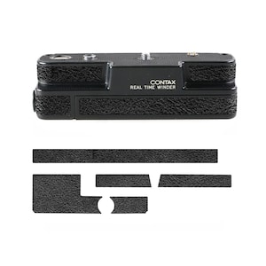 Puede incluir: Accesorio de cámara Contax Real Time Winder negro con agarre texturizado y el texto "CONTAX REAL TIME WINDER". Incluye varias almohadillas adhesivas negras para su aplicación. El accesorio de la cámara se muestra sobre un fondo blanco.