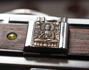 May include: Close-up of a camera with a detailed metal Buddha figure on the latch. The camera body features a dark wood grain finish and a silver metal frame. A small, yellow circular detail is visible.