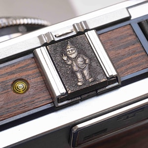 Può includere: Primo piano di una fotocamera con un design argento e venature del legno. Una placca di metallo con una figura di gnomo che fa il dito medio è attaccata alla fotocamera. Lo gnomo indossa un cappello e una giacca.