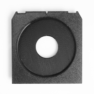 May include: A black, square-shaped object with a circular opening in the center. The object has a textured surface and a smaller, white circular hole in the middle. The corners of the square have small cutouts.