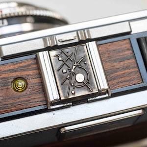 May include: Close-up of a vintage camera with a silver body and wooden accents. The camera features a decorative metal plate with an abstract design. A small, yellow bubble level is visible on the wooden section.