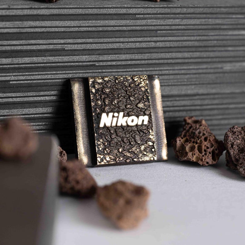 May include: A square metal camera hot shoe cover featuring a textured, dark, rugged surface with the word Nikon printed in white lettering. The accessory has smooth, metallic side edges and is surrounded by small, porous, dark brown volcanic rocks.
