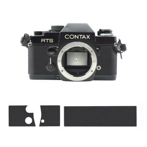 Puede incluir: Una cámara Contax RTS negra con una montura de lente plateada y la palabra "CONTAX" impresa en la parte superior. La cámara tiene una correa negra y la palabra "RTS" está impresa en el lateral. Debajo, tres almohadillas negras para el agarre de la cámara.
