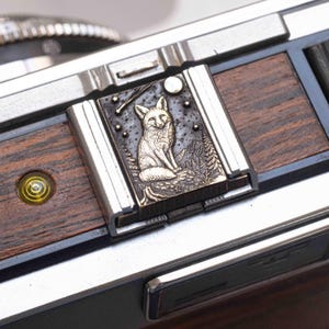 Puede incluir: Una tapa de zapata metálica rectangular para cámara, con un diseño en relieve de un zorro sentado en un bosque bajo la luna y las estrellas. El accesorio está montado en un cuerpo de cámara con paneles de madera oscura y un nivel de burbuja.