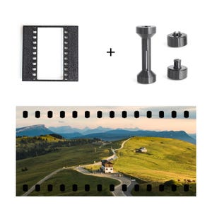 May include: A black rectangular film frame holder, a central spool, and two circular end caps are displayed alongside a panoramic landscape photograph featuring a winding road through green hills, mountains, and a building under a soft sky.