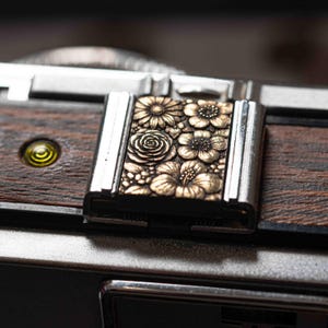 Puede incluir: Primer plano de una cámara vintage con un diseño floral detallado. La cámara presenta un acabado de madera oscura y una placa metálica decorativa con flores en relieve. Los detalles metálicos son plateados y dorados.