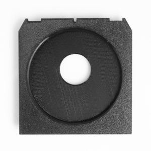 May include: A black, square-shaped object with a circular indentation in the center. The center has a hole. The object has a textured surface and appears to be made of a solid material. The design includes a square frame with a circular inset.