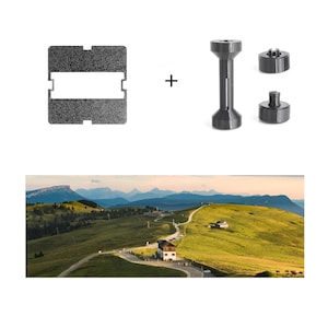 Könnte beinhalten: Eine schwarze, texturierte rechteckige Platte mit einem mittigen Ausschnitt, eine schwarze zylindrische Spule und zwei kleine schwarze kreisförmige Komponenten. Darunter befindet sich ein Landschaftsfoto einer kurvigen Straße durch grüne Hügel und Berge.