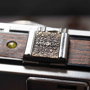 May include: Close-up of a vintage Minolta camera. The camera features a textured metal plate with the brand name, set against a backdrop of wood grain and silver metal. A small, yellow-green light is visible on the side.