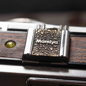 Pode incluir: Close-up de uma câmera Mamiya vintage. A câmera apresenta uma placa texturizada de cor bronze com o nome da marca "Mamiya" em letras brancas. O corpo da câmera tem um acabamento em grão de madeira e detalhes prateados.