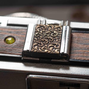 May include: Close-up of a vintage camera with an intricate floral pattern on the top. The camera has a dark wood finish and silver metal detailing. A small, yellow-tinted lens is visible on the side.