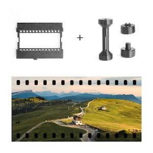 Könnte beinhalten: Ein Satz dunkelgrauer 3D-gedruckter Filmentwicklungswerkzeuge und ein Filmstreifen mit einer malerischen Landschaft. Die Werkzeuge umfassen einen Filmhalter, einen zentralen Pfosten und zwei kleinere Komponenten. Der Filmstreifen zeigt eine kurvenreiche Straße durch grüne Hügel und Berge.