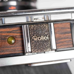 Peut inclure: Gros plan sur un appareil photo Rollei vintage. L'appareil photo présente un corps en métal argenté avec une plaque marron et dorée texturée. Le mot "Rollei" est en relief sur la plaque. L'appareil photo a un panneau en bois avec une lentille jaune.
