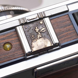 May include: Close-up of a camera with a decorative metal plate featuring an illustration of The Little Prince. The plate is bronze-coloured with a detailed engraving of the character, a rose, stars, and a moon. The camera has a wooden and metallic finish.