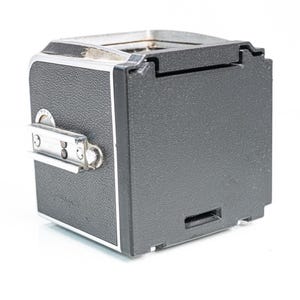 Könnte beinhalten: Eine Vintage-Kamera in Schwarz und Silber mit strukturiertem Gehäuse. Die Kamera hat eine rechteckige Form mit silberner Zierleiste und Blitzschuh. Ein klassisches Stück Fotoausrüstung.