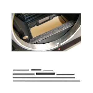 Op de afbeelding: Close-up van de binnenkant van een camera, met een zilveren metalen frame en een zwarte, getextureerde schuimstrip. De binnenkant toont een lichtbruin oppervlak, met een kleine rode stip aan de zijkant van het frame. De afbeelding benadrukt de interne componenten van de camera.