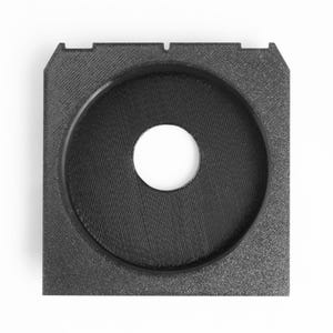 May include: A square, black, 3D-printed object with a circular indentation and a central hole. The object has a textured surface and a raised outer edge. The central hole is white, contrasting with the black material.