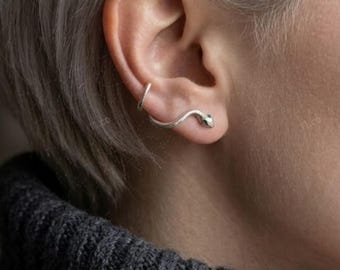 May include: A silver ear cuff and stud earring set. The ear cuff curves along the ear, and the stud is a small snake head. The person is wearing a dark gray turtleneck sweater.