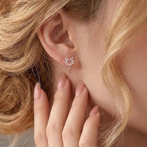May include: A close-up of a person's ear wearing a pair of silver fox-shaped earrings. The earrings have a fox head design with a dangling tail. The person has blonde hair and is wearing the earrings on their left ear.