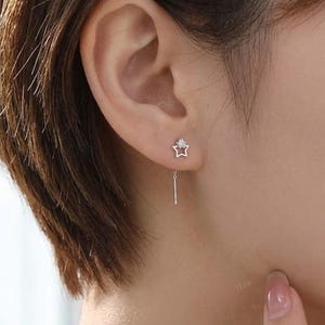 Puede incluir: Primer plano de un pendiente plateado con un perno en forma de estrella y una barra colgante. La estrella tiene una pequeña gema transparente en el centro. El pendiente se lleva en la oreja de una persona con pelo corto y castaño.