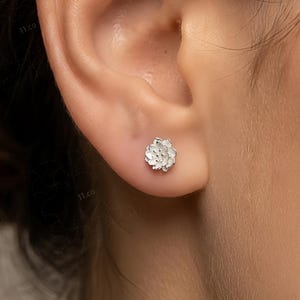 May include: A silver flower-shaped stud earring is shown on an ear. The earring has a detailed, textured design, resembling a small, delicate flower. The background is a blurred view of skin.