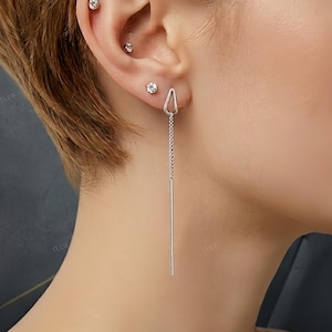 May include: A close-up of a person's ear with multiple piercings. The ear features three small, round, clear stud earrings and a long, dangling silver earring with a triangular top. The background is a dark, textured gray.