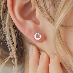 May include: A close-up of an ear with a small, silver stud earring. The earring has a minimalist design, a hollow triangle shape. The earring is set against the backdrop of the person's skin and hair.