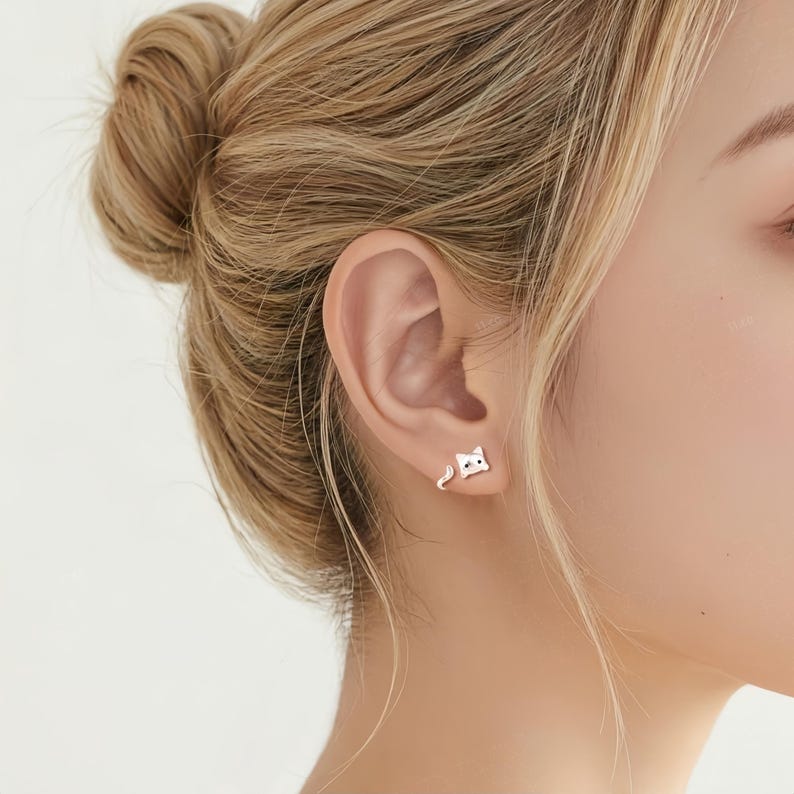 May include: A silver-toned stud earring shaped like a cat, featuring a small head with two black eyes and a separate curved tail piece that wraps around the earlobe. The design is minimalist, metallic, and polished, creating a two-part cat figure.