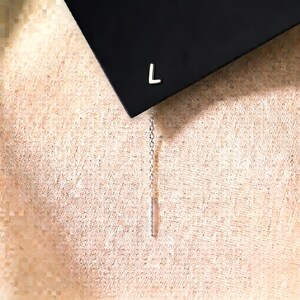 May include: A silver-toned earring featuring a small, capital letter L stud attached to a delicate, thin chain that hangs downwards, ending in a slender, straight metal bar. The earring is displayed against a dark, angled surface.