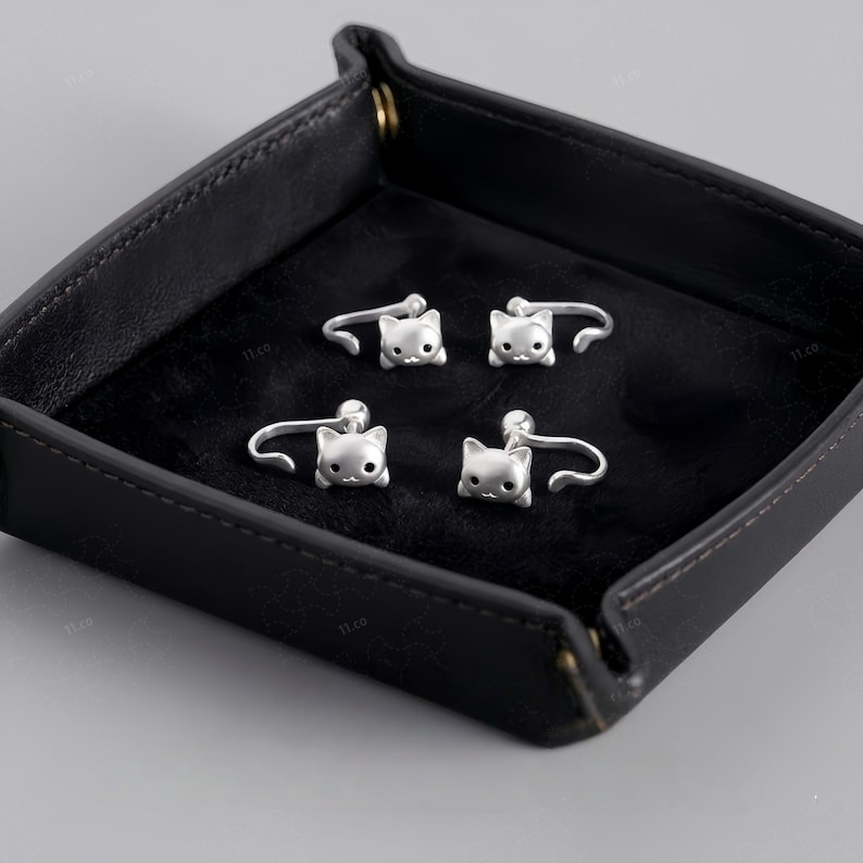 May include: Four silver-toned ear cuffs, each featuring a small cat head design with pointed ears and dark eyes. The cat heads are smooth and metallic, attached to a curved band that wraps around the ear cartilage. These jewelry pieces are arranged in a square formation, showcasing the detailed feline facial features and the polished finish of the metal construction.