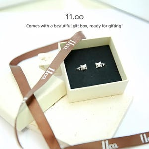 May include: A pair of small white cat-shaped stud earrings resting inside a square cream-colored gift box with a black velvet insert. A brown ribbon with the text 11.co printed in white is draped around the open box, which is labeled 11.co.