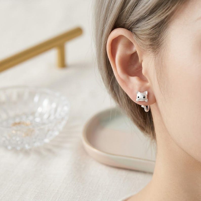 May include: A silver-toned stud earring shaped like a cat head with small ears and black eyes. The design features a curved tail element that extends from the back of the earlobe, creating a wrap-around effect that mimics a cat clinging to the ear.