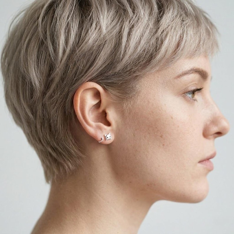 May include: A silver-toned stud earring shaped like a small cat head with two pointed ears and two dark eyes. The earring is positioned on the earlobe, featuring a curved silver wire extending from the base of the cat head along the lower earlobe.