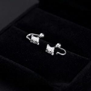 May include: A pair of silver-toned earrings featuring a cat head design. Each earring has a curved post that extends from the back of the cat head, ending in a small spherical ball closure. The cat faces are smooth with small dark eyes.