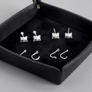 May include: A set of silver-toned stud earrings featuring cat head designs with small black eyes, paired with separate silver-toned curved ear cuffs that resemble cat tails, all displayed on a black velvet-lined square tray.