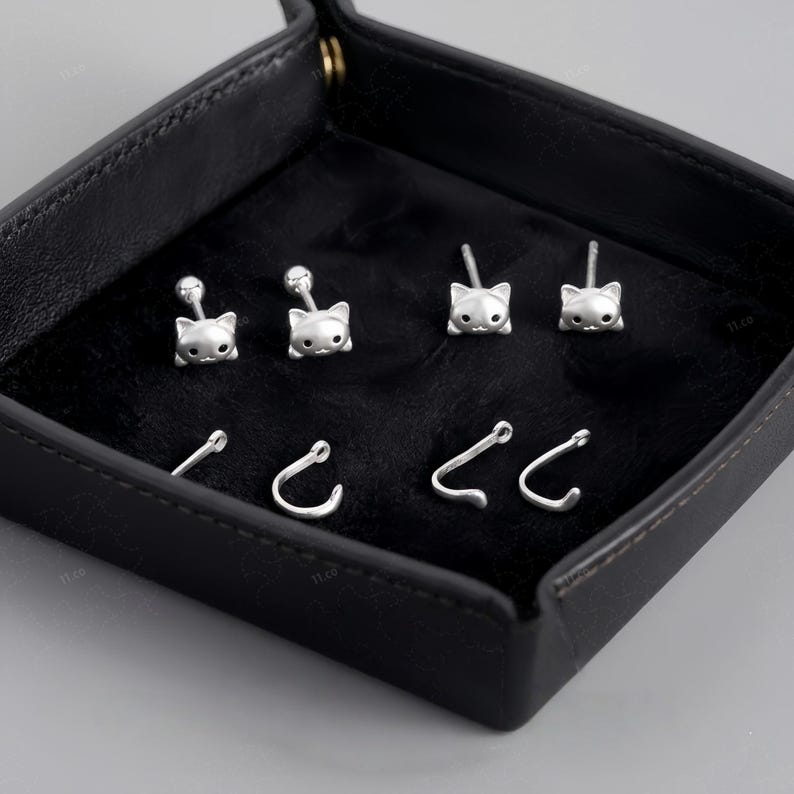 May include: A set of silver-toned stud earrings featuring cat head designs with small black eyes, paired with separate silver-toned curved ear cuffs that resemble cat tails, all displayed on a black velvet-lined square tray.