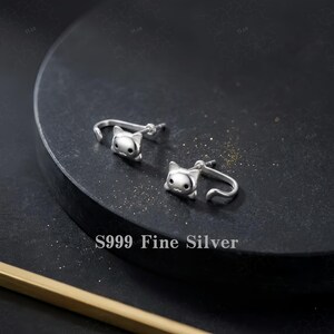 May include: A pair of S999 fine silver stud earrings shaped like cat heads with small ears and black eyes. The earrings feature a curved post design that wraps slightly around the earlobe, crafted from polished silver with a smooth, reflective finish.