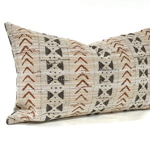 May include: A rectangular decorative pillow with a beige and brown geometric pattern. The pillow features a repeating design of arrows and bow tie shapes in shades of brown and black. The pillow is made of a textured fabric.