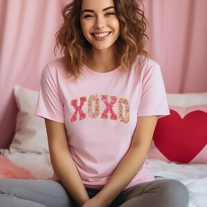 May include: A light pink t-shirt with the text "XOXO" in a glittery pink and gold design. The shirt is short-sleeved and appears to be made of a soft material. The image is of a person smiling.