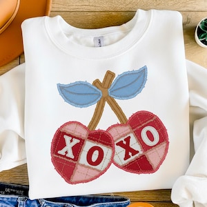 Può includere: Felpa bianca con un design a ciliegia. Ogni ciliegia è rossa e rosa con le lettere "XOXO". Il design include foglie blu e steli marroni. La felpa è del marchio Gildan.