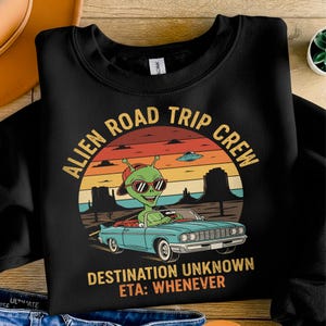Puede incluir: Sudadera negra con un gráfico de un extraterrestre conduciendo un descapotable azul. El diseño incluye el texto "Alien Road Trip Crew" y "Destination Unknown ETA: Whenever" en un diseño retro de puesta de sol.