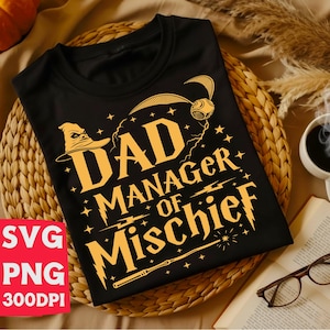 May include: Black t-shirt with a gold graphic that reads "DAD MANAGER OF MISCHIEF" with a wizard hat, lightning bolts, and a wand. The shirt is displayed on a woven basket with a book, glasses, and a cup of coffee.